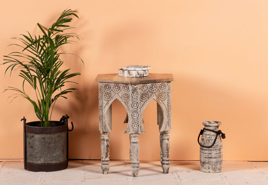 Decorative table with intricate carvings next to a potted plant and a vase against a peach-colored wall.