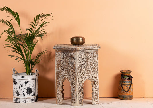 Decorative table with intricate carvings next to a plant and a vase against a peach-colored wall.